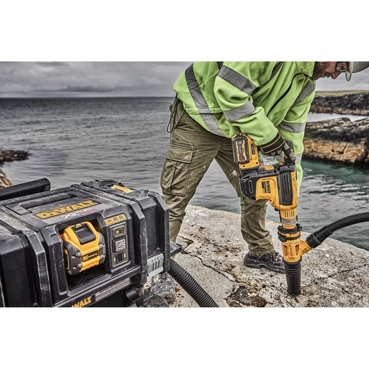 A person in a high-visibility jacket operates a DEWALT power tool to drill into a stone surface outdoors near water, with a DEWALT tool case and battery pack in the foreground.