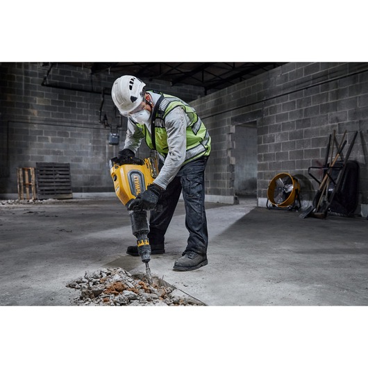 A person wearing a safety helmet, high-visibility vest, gloves, and a dust mask is using a DEWALT power tool to break concrete on the floor inside an industrial building.