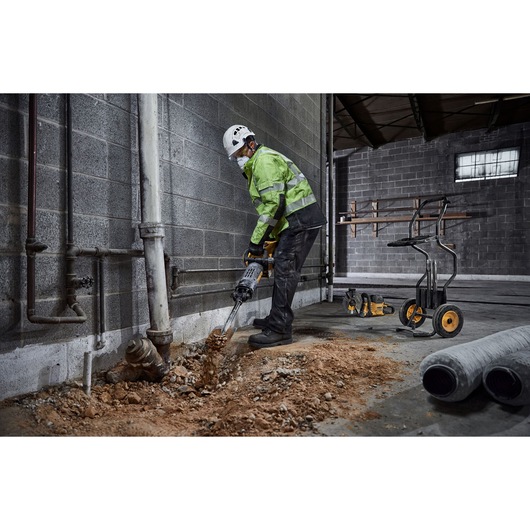 A person wearing safety gear uses a DEWALT demolition hammer to break up a concrete floor near a wall in an industrial setting. Construction tools and equipment are visible in the background.