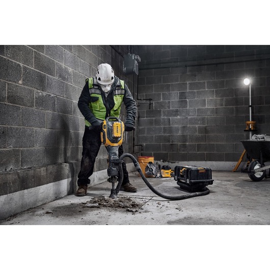 A construction worker in safety gear operates a DEWALT electric jackhammer to break up concrete on a worksite. The tool is connected to a vacuum system and there is construction equipment in the background.
