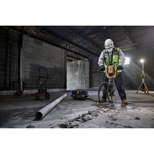 A construction worker wearing a safety helmet and reflective vest uses a DEWALT demolition tool to break concrete in an industrial setting. Nearby are a pipe, a tool case, a dolly, and a standing work light.