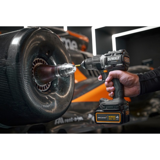 A person using a DEWALT cordless drill on the wheel of a racing car. The drill is labeled with McLaren and DEWALT branding, and the car in the background features orange and black colors.