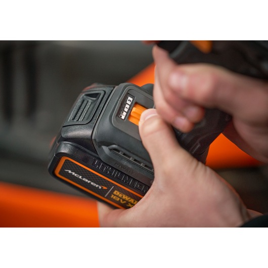 Close-up of hands holding a DEWALT 18V lithium-ion battery pack, showing the battery status indicator and part of an orange power tool in the background.