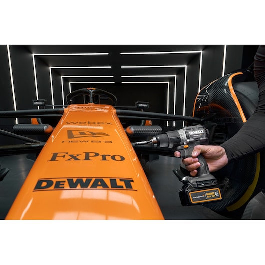 A person uses a DEWALT cordless drill on the front suspension of an orange Formula 1 racing car with visible sponsor logos including DEWALT, FxPro, New Era, and Webex. The scene is set in a modern, futuristic garage with illuminated lines on the walls and ceiling.