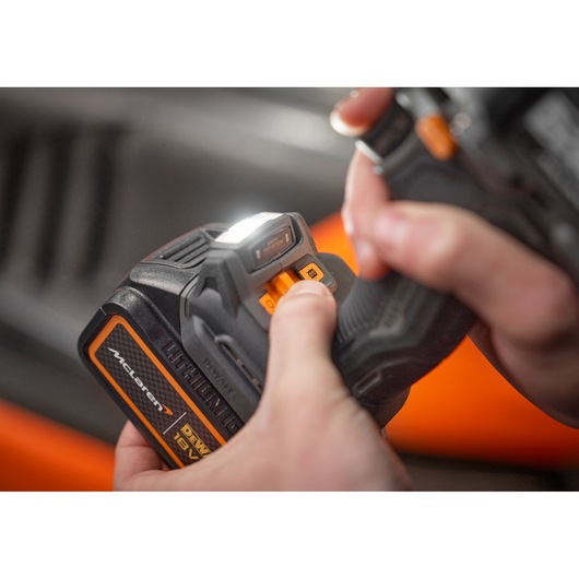 Close-up of a person holding a DEWALT cordless power tool with a McLaren 18V battery. The person's thumb is pressing an orange button and a white LED light on the tool is illuminated.
