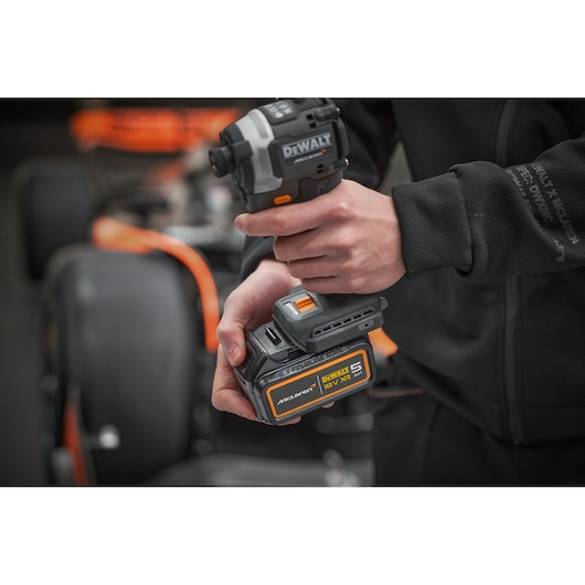 A person holding a DEWALT cordless power tool with an 18V XR lithium-ion battery, standing in front of a vehicle with orange and black details.