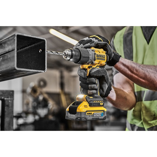 A close-up of a person wearing gloves and a high-visibility vest, using a yellow DEWALT cordless drill to drill into a metal pipe in an industrial workshop.