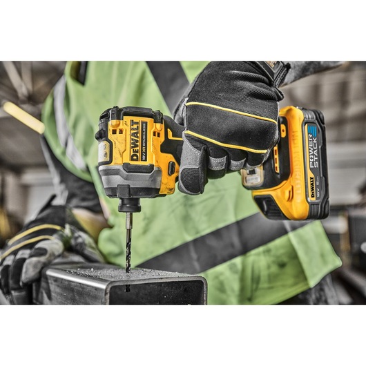 A person wearing work gloves and a green safety vest is using a DEWALT cordless drill to drill into a metal surface.