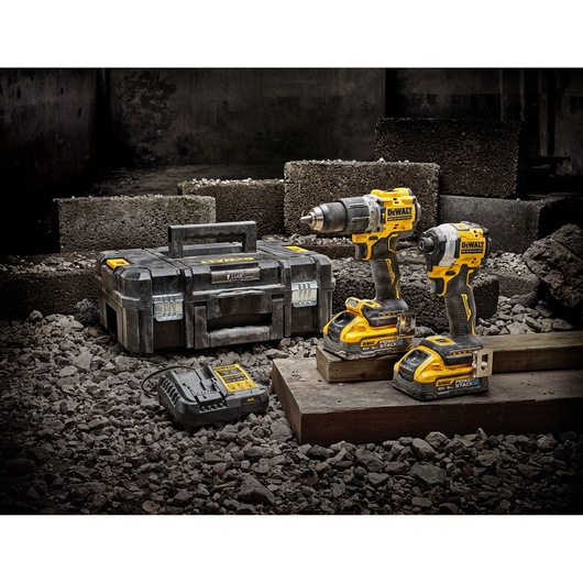 DEWALT power tool kit including two cordless drill drivers, a battery charger, and a sturdy black and yellow carrying case, shown on a construction site with concrete blocks and gravel.