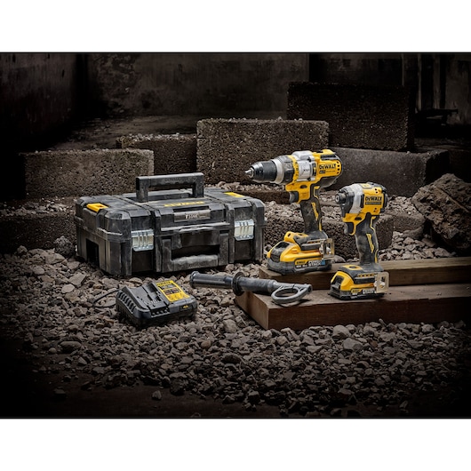 A DEWALT tool set displayed on a rocky surface, featuring a large black and yellow toolbox, a cordless drill, an impact driver, a battery charger, a battery pack, and a drill handle. The background includes concrete blocks and construction debris.