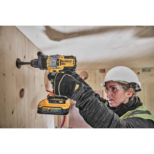 A person wearing a hard hat and gloves uses a DEWALT cordless drill to drill a hole in a wooden wall. The DEWALT Power Stack 18V battery is attached to the drill. The person's face is blurred for privacy.