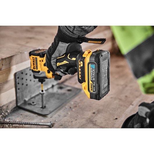 A person wearing protective gloves uses a yellow and black DEWALT cordless power drill with a POWERSTACK battery to drive a screw into a metal bracket attached to a wooden beam at a construction site.