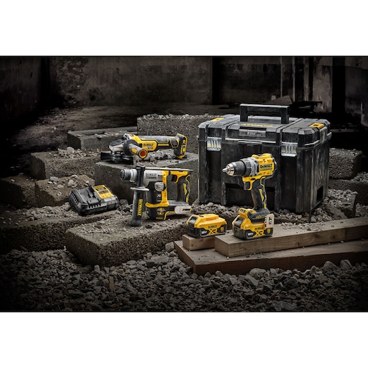A set of DEWALT power tools including a cordless drill, an angle grinder, a rotary hammer, batteries, a charger, and a large black tool case, displayed on concrete blocks and gravel in an industrial setting.