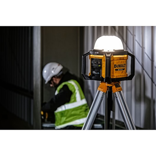 DEWALT DCL074 LED work light on tripod, used at construction site with worker in safety gear in background.