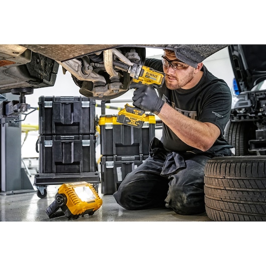 Person using DEWALT tools on car suspension, with DEWALT work light on floor and tool storage boxes nearby.
