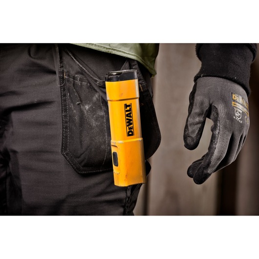 A close-up of a person wearing work gloves and carrying a yellow DEWALT portable light in the pocket of black work trousers.