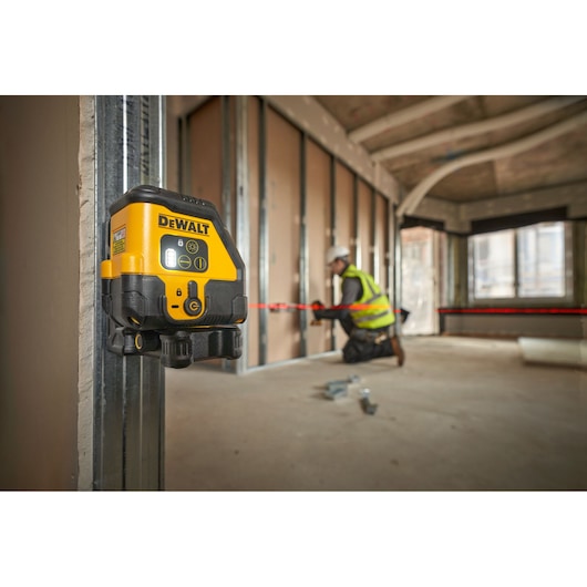 DEWALT laser level tool attached to a metal stud in a construction site, showing red laser lines across the unfinished interior walls. A person wearing a safety vest and hard hat is working in the background.