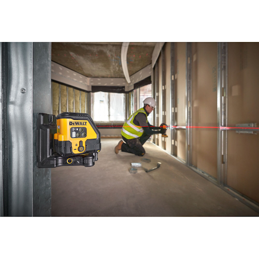 A DEWALT laser level device mounted on a metal frame inside a room under construction, with a worker in a safety vest and hard hat using the red laser line to measure or align materials along a wall.