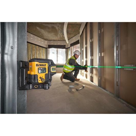 A DEWALT laser level device mounted on a metal beam emits a bright green laser line across a construction site, where a person wearing a safety vest and helmet is working with tools near a wall frame.