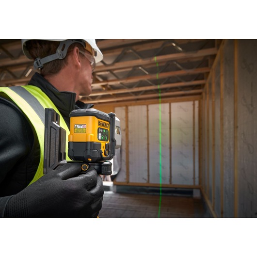 A person wearing safety gear uses a DEWALT laser level tool inside a wooden-frame construction site. The tool is projecting a green laser line onto the wall.