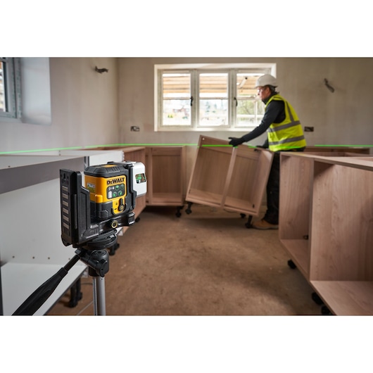 A DEWALT laser level device mounted on a tripod emits a green laser line across the room. In the background, a construction worker wearing a safety vest and helmet is installing wooden cabinets. The scene appears to be inside a partially completed room with a window and unfinished walls.
