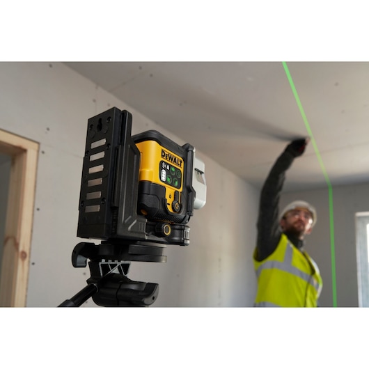 A DEWALT laser level device mounted on a tripod projects a bright green line onto a ceiling in a construction area. A person wearing a high-visibility vest is marking or measuring along the line.