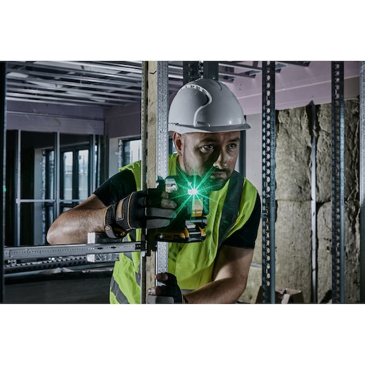 A person wearing a white hard hat, high-visibility vest, and gloves is using a DEWALT laser level tool to align a metal stud in a construction environment. The green laser is activated and visible in the image.