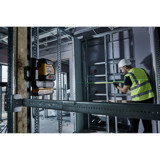 A DEWALT laser level device (model DCLE34021) is mounted on a wooden beam in a construction site. A person wearing a high-visibility vest and a white hard hat is measuring in the background.
