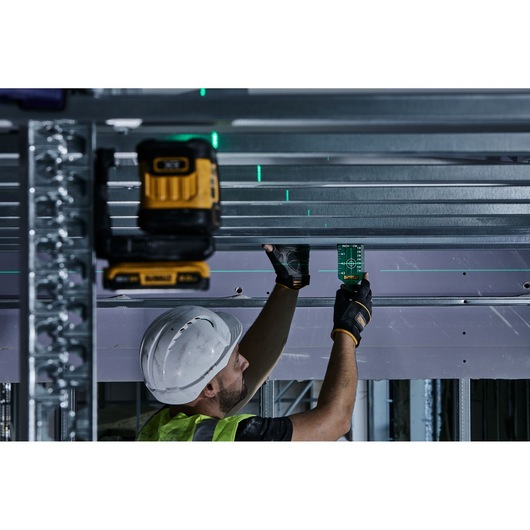 A worker wearing a hard hat and gloves is using a DEWALT laser level tool on a construction site, aligning metal beams on a ceiling. The worker holds a DEWALT green laser target, surrounded by structural materials.