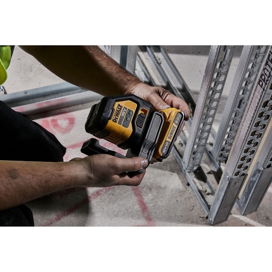 A person holding a DEWALT cordless laser level tool, model DCLE34021, in a construction environment near metal framing and a concrete floor.