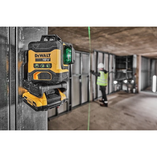 A DEWALT 18V laser level device mounted on a metal surface in a construction site, projecting a green laser line. In the background, a person wearing a safety vest and helmet is working with metal wall frames.