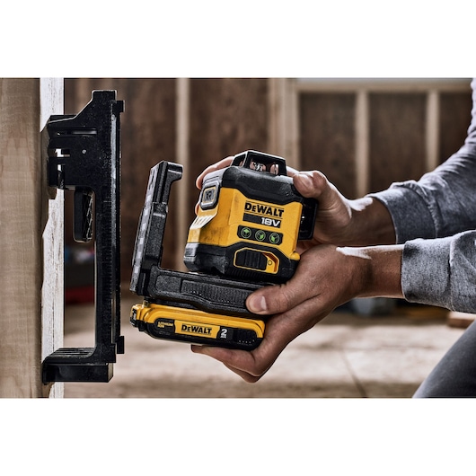 A person holding a DEWALT 18V laser level device against a wall, with a mounting bracket attached. The device is yellow and black, used for precise measurements in construction or carpentry.