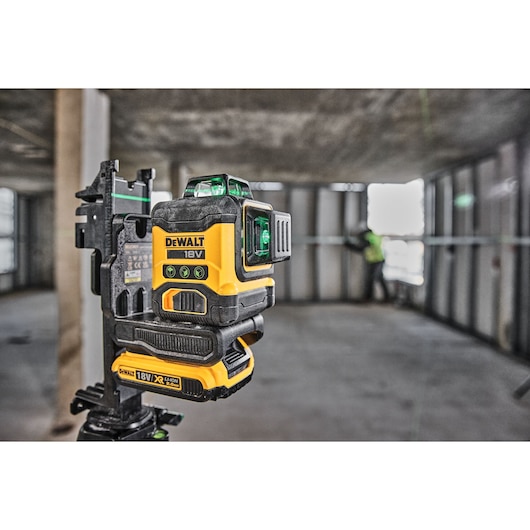 A DEWALT 18V green line laser level (SKU: DCLE34031D1) is positioned on a tripod at a construction site. The tool is in use, projecting green laser lines onto unfinished walls, with a worker visible in the background.