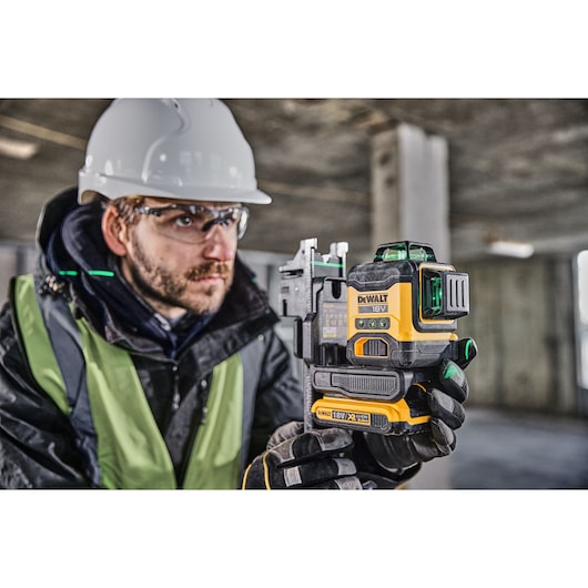 A person wearing a safety helmet and high-visibility vest holds a DEWALT 18V green laser level tool inside a construction site.