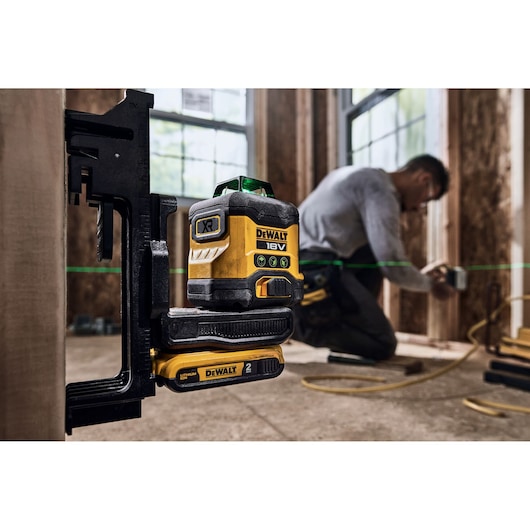A DEWALT 18V XR green laser level, model DCLE34031N, is mounted on a clamp in a construction setting. A person is working in the background, adjusting something on a wooden frame, while green laser lines are visible, indicating alignment.