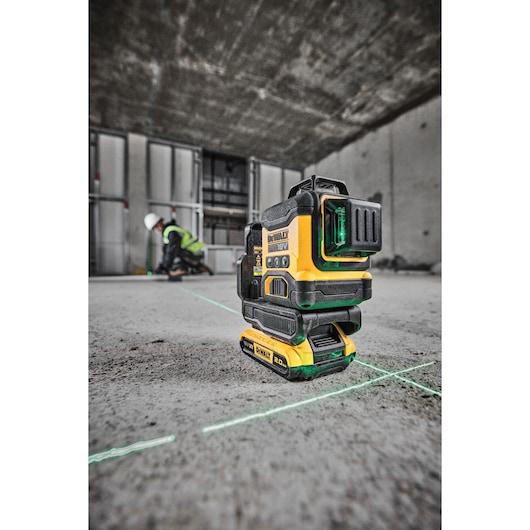 A DEWALT 18V green laser level tool is placed on a concrete floor, projecting bright green laser lines. In the background, an individual wearing a safety helmet and vest works in an unfinished construction site with exposed metal framing.