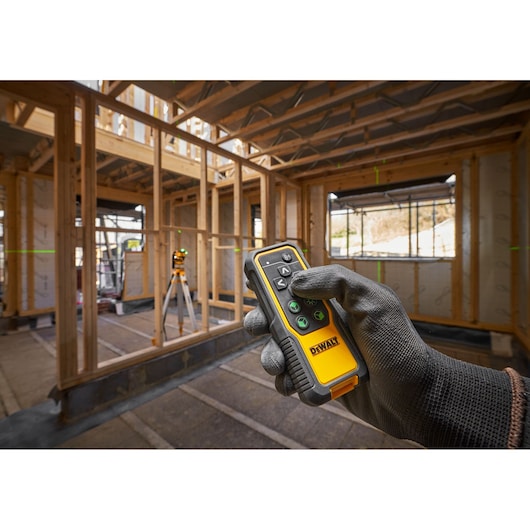 A gloved hand holding a DEWALT remote control device in the foreground, with a DEWALT laser level positioned on a tripod in a wooden house frame construction site in the background. Green laser lines are visible across the site, aiding alignment and measurements.