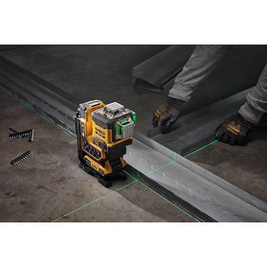 A DEWALT laser level tool projecting green laser lines on a construction site floor, with a person wearing gloves positioning metal beams along the lines.