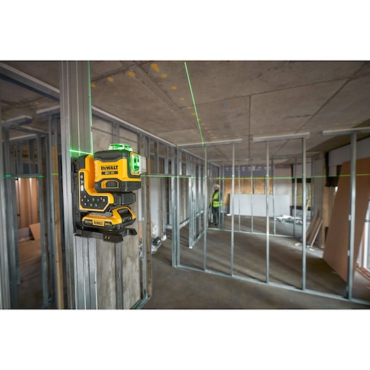 A DEWALT laser level is mounted on a metal stud in a construction site, projecting visible green laser lines across the walls and ceiling framing during building layout work.