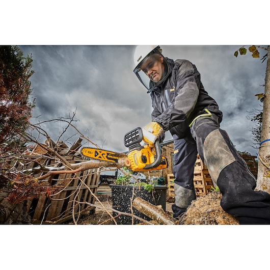 Person wearing safety gear uses a DEWALT chainsaw to cut branches outdoors on a cloudy day.