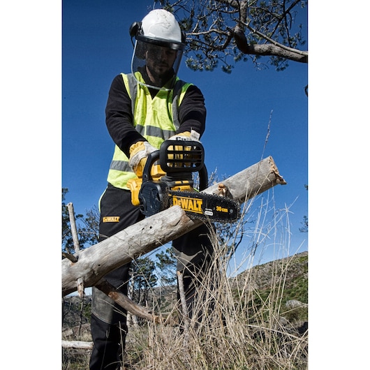 Person in safety gear using a DEWALT DCM565N chainsaw to cut a log outdoors. Clear blue sky in the background.
