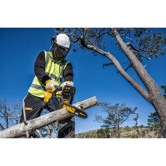 Worker using a DEWALT chainsaw to cut a tree branch outdoors. Safety gear and clear sky are visible.