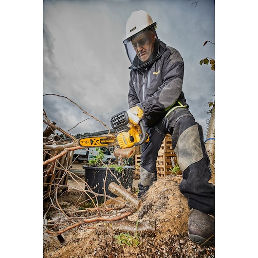 Person uses DEWALT chainsaw DCM565N to cut tree branches outdoors, wearing safety gear.