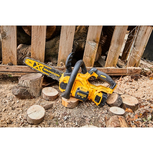 Yellow DEWALT cordless chainsaw DCM565N on ground, surrounded by wood logs and sawdust outdoors.