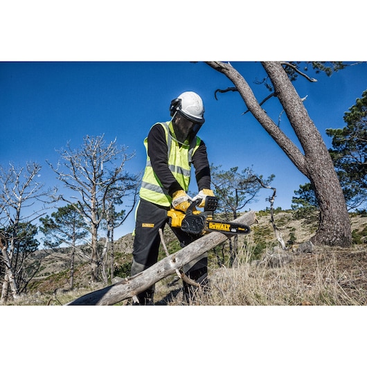 Person using a DEWALT DCM565N chainsaw to cut a tree branch outdoors, wearing safety gear and a vest.