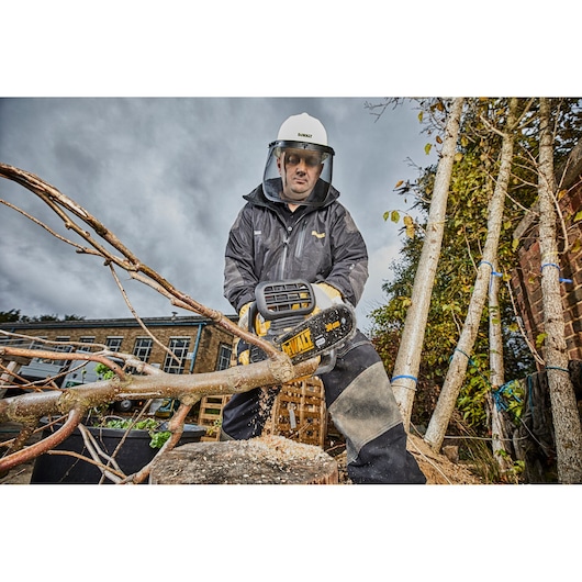 Person using DEWALT DCM565 chainsaw to cut tree branches outdoors, wearing safety gear.