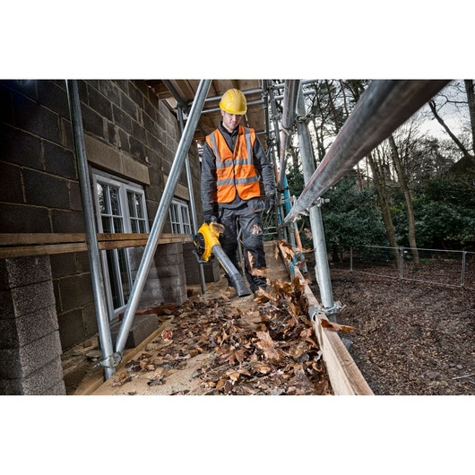 Worker uses a DEWALT leaf blower to clear leaves from scaffolding at a construction site.