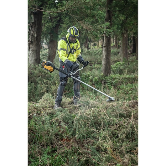 A person wearing high-visibility protective work clothing operates a DEWALT brush cutter on a grassy, uneven terrain in a wooded area.
