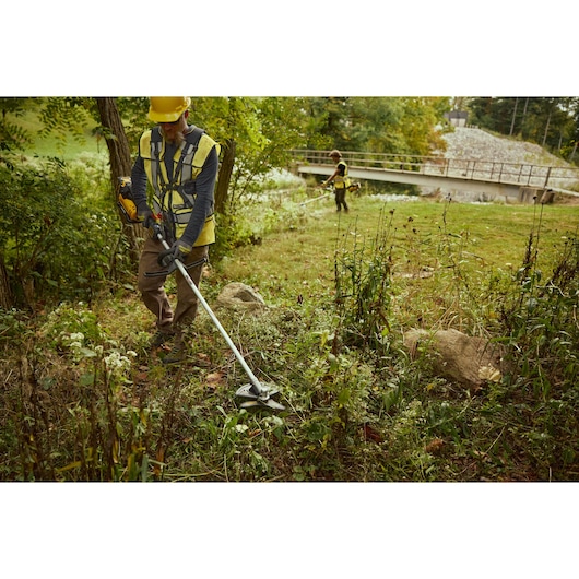Two workers wearing safety vests and helmets are using DEWALT brush cutters to trim overgrown vegetation in an outdoor area near a bridge.