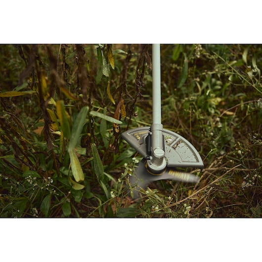 Close-up of a DEWALT brush cutter in use, trimming grass and weeds outdoors.